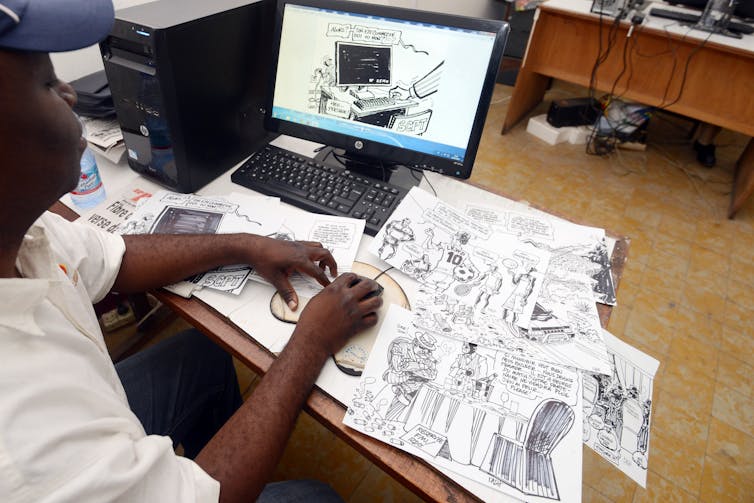 A man sits at a desk that's covered in hand drawn cartoons, touching one up on a computer screen in front of him.