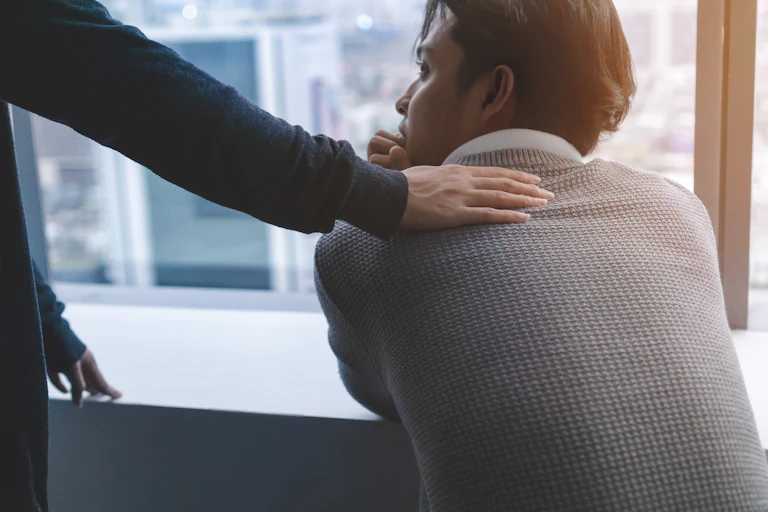 Man stares out window as person behind touches his back