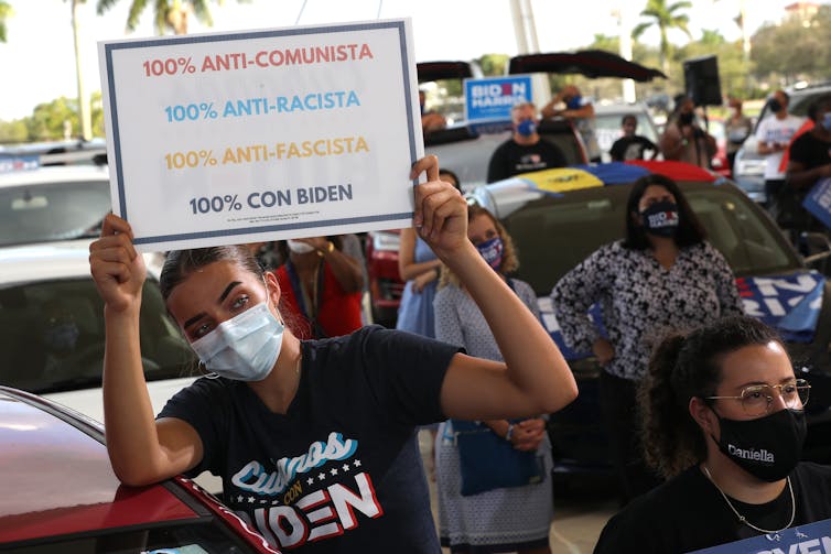Woman in a 'Cubans for Biden' shirt holds a sign reading '100% anti-communism, 100% pro-Biden'