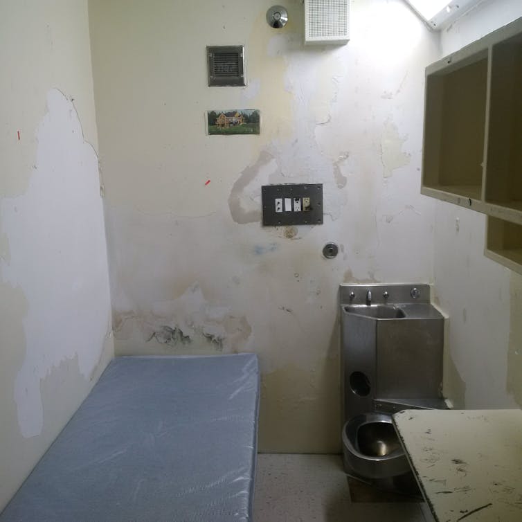 A small prison cell with a cot, sink, toilet and desk