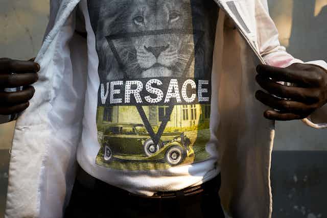 The middle section of a man's body, dressed in a T-shirt with a spangled VERSACE logo, revealed by two hands holding open a shiny white jacket.