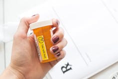 A woman's hand holding a prescription bottle with a prescription pad in the background.