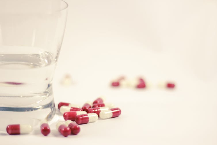 Red and white pill capsules scattered on a white surface beside a glass of water.