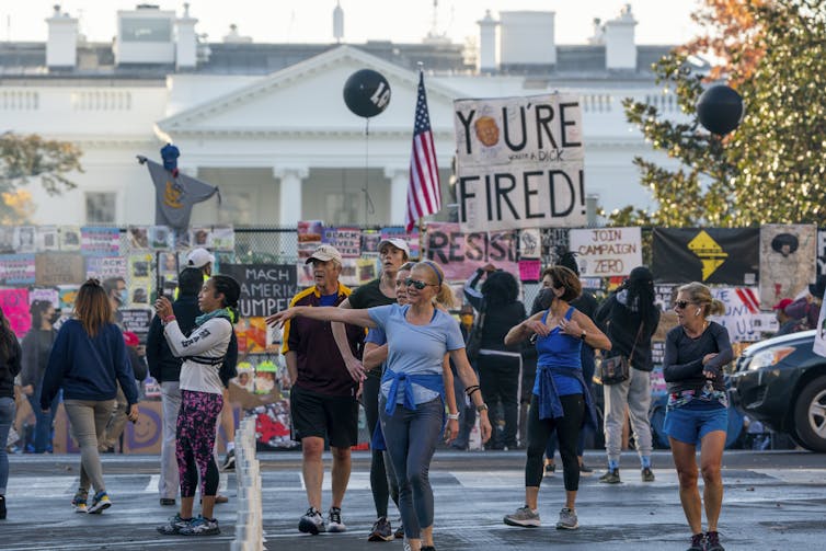 People carrying anti-Trump signs gather with the White House seen in the background.