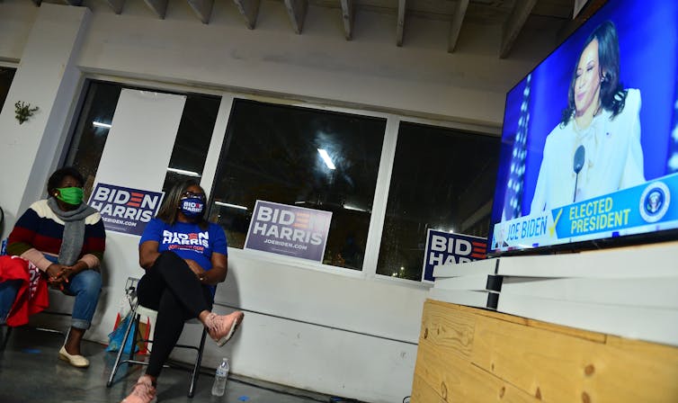 Two Black women wearing Biden/Harris shirts watch Harris, in white, speak on TV