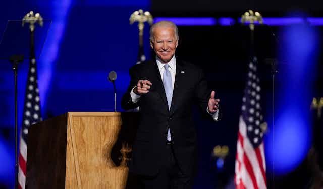 Joe Biden smiles and points to someone in the crowd from a stage with American flags behind him.
