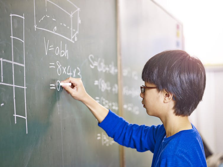 A young Asian boy writing calculations on a chalk board.