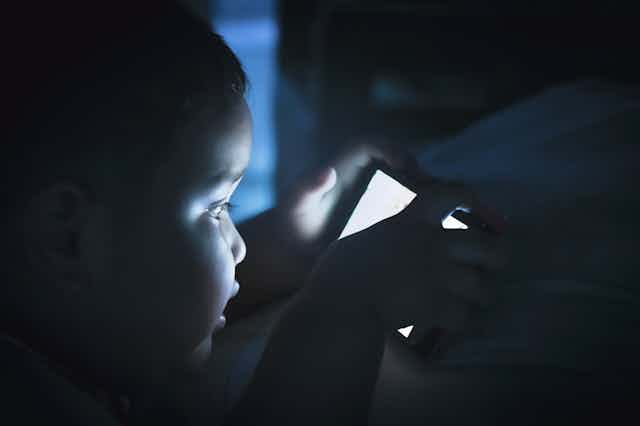 Un niño pequeño a oscuras con el rostro iluminado por la pantalla de un teléfono móvil.