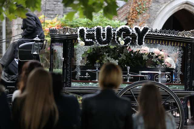 A horse-drawn hearse decorated with the name 'Lucy' carries a coffin.