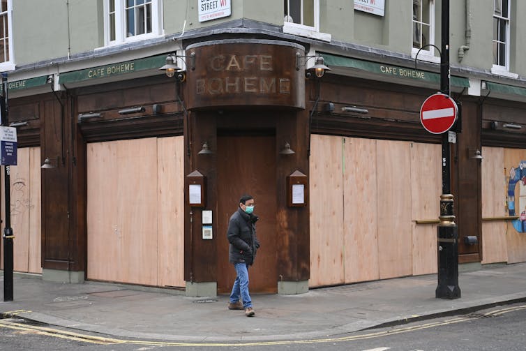Man wearing a face mask walks past a boarded up cafe.