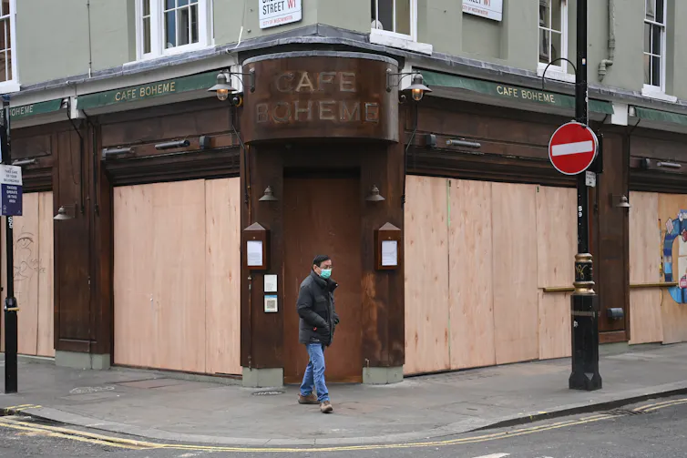 Man wearing a face mask walks past a boarded up cafe.
