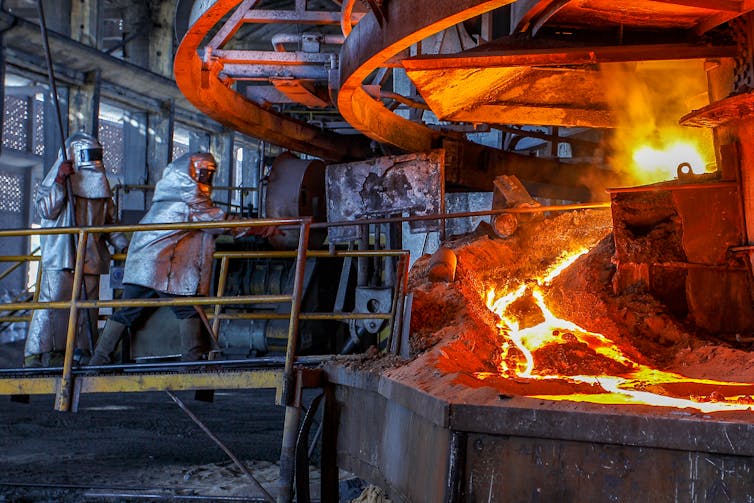 A worker in silver, protective gear stokes a furnace spewing molten metal.