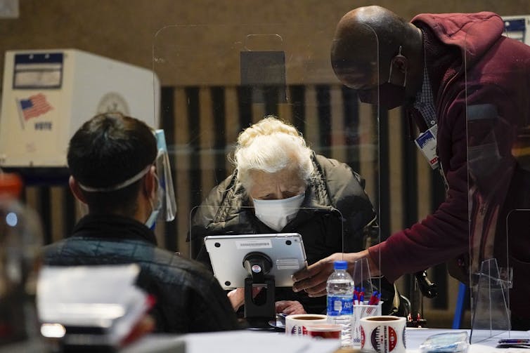 A white-haired woman signs in to vote as two poll workers provide guidance.