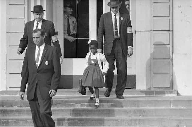 Men in suits escort a little girl in a dress outside of a building in an old photo..