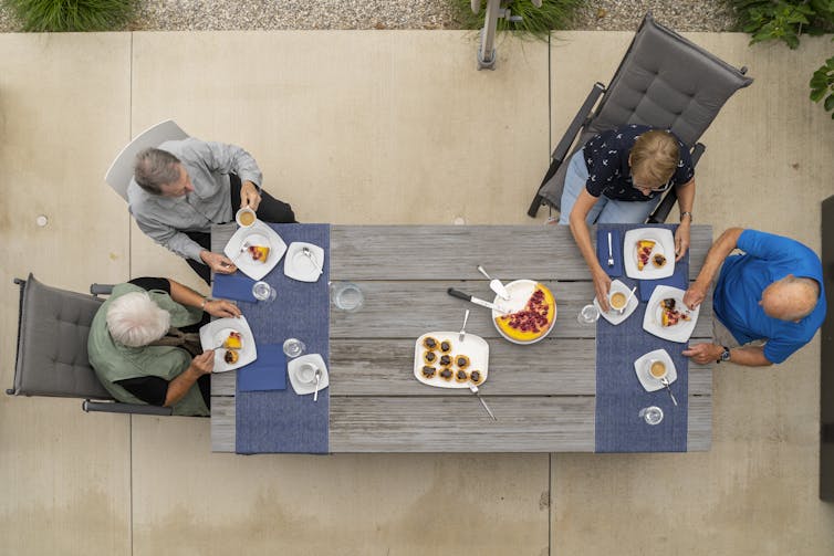 Two couples sit at opposite ends of a table outside while eating a meal.