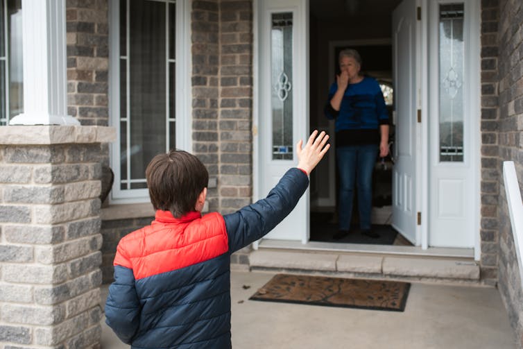 A young boy waves to his grandmother at the door of a home.