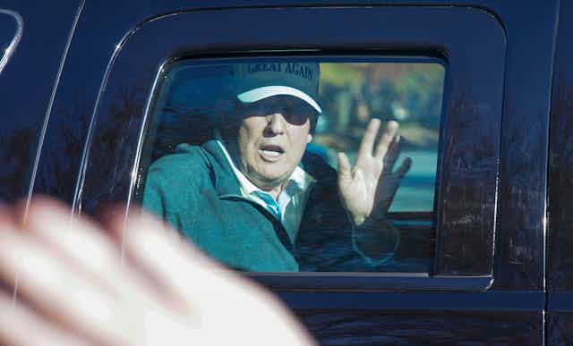 Trump waves from the back of a limousine wearing a white Make America Great Again ball cap.