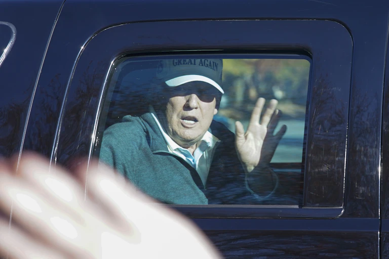 Trump waves from the back of a limousine wearing a white Make America Great Again ball cap.