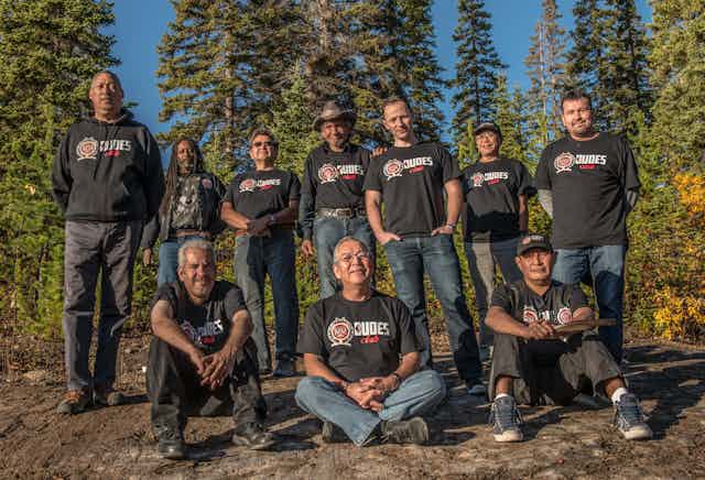 A group of 10 men — three sitting on the ground and seven standing behind them — wearing DUDES Club shirts outdoors in front of large evergreen trees.