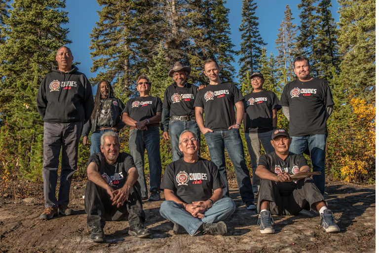 A group of 10 men — three sitting on the ground and seven standing behind them — wearing DUDES Club shirts outdoors in front of large evergreen trees.