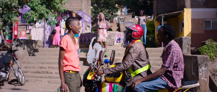 An urban street scene where two men on a stationary motorbike chat to a young woman standing in front of them and another young woman with pink hair extensions comes down some stairs towards them in the background.