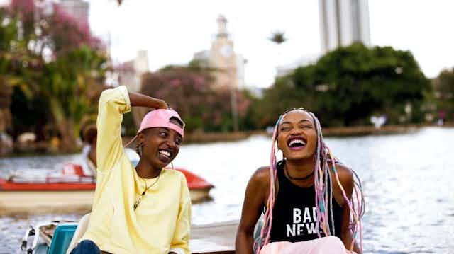 Two young women in a boat on a lake in the city. One, with pink hair extensions, is laughing delightedly and the other, an arm on her head adorned with a pink backward peak cap, beams.