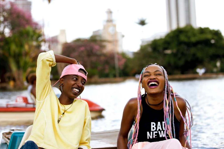 Two young women in a boat on a lake in the city. One, with pink hair extensions, is laughing delightedly and the other, an arm on her head adorned with a pink backward peak cap, beams.