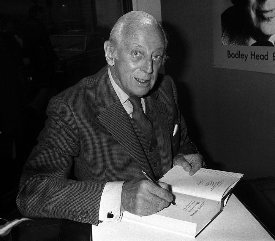 Elderly Alistair Cooke in a suit signing a book.