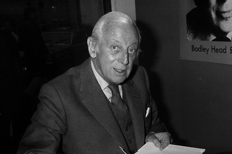 Elderly Alistair Cooke in a suit signing a book.