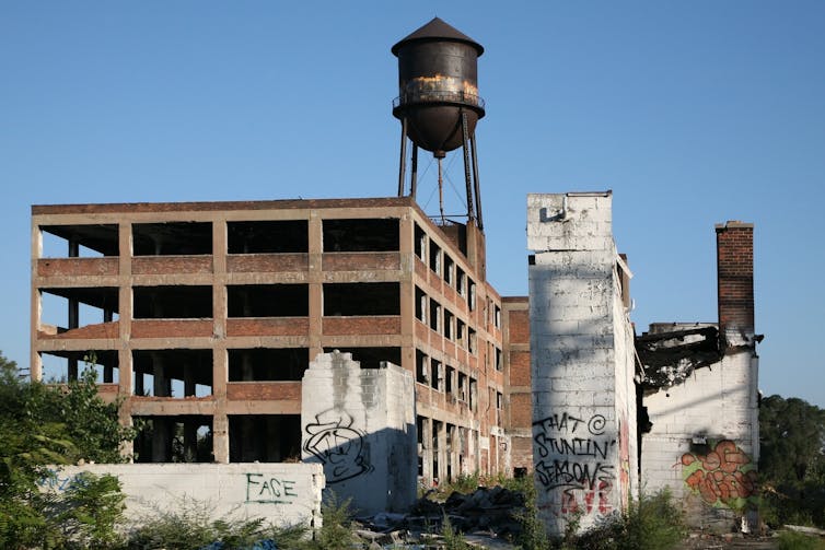 An abandoned factory sits amid overgrown vegetation, with a rusted water tower behind it.