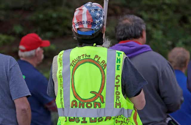 A group of people face away from the camera. A man in the foreground wears a Stars & Stripes cap and a florescent safety vest with writing supporting the Qanon conspiracy theory in hand lettering.