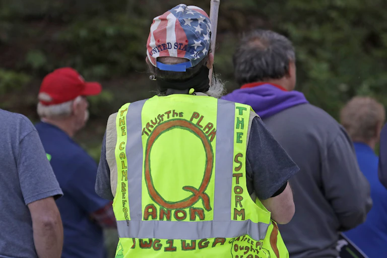 A group of people face away from the camera. A man in the foreground wears a Stars & Stripes cap and a florescent safety vest with writing supporting the Qanon conspiracy theory in hand lettering.