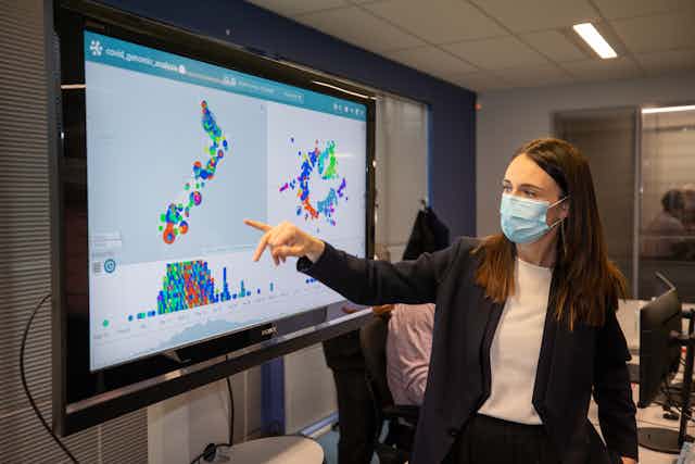 Jacinda Ardern wearing a mask, pointing at a map of New Zealand