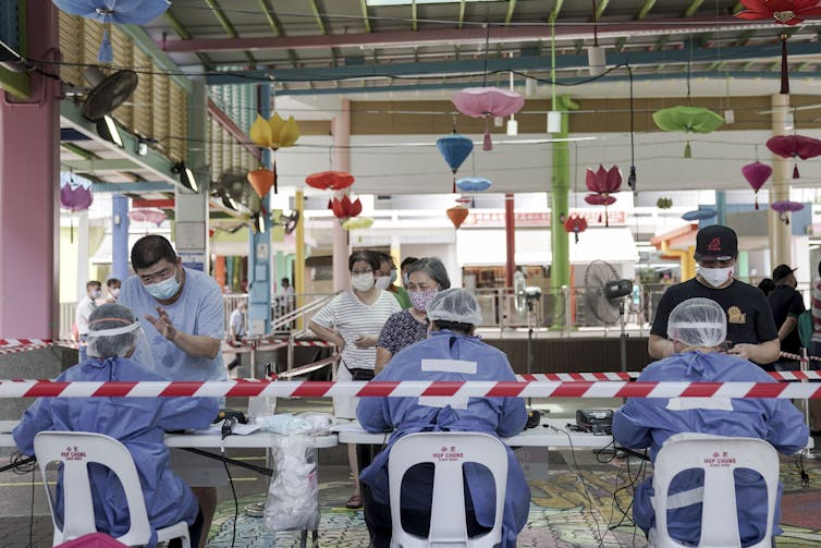 Workers in PPE register people at a table with colourful lights hanging behind.