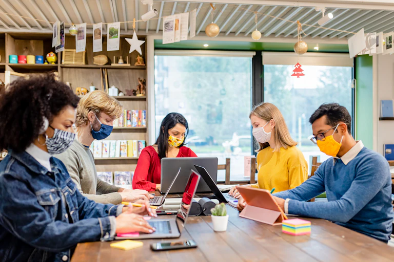 Five people wearing facemasks sat around desk working.