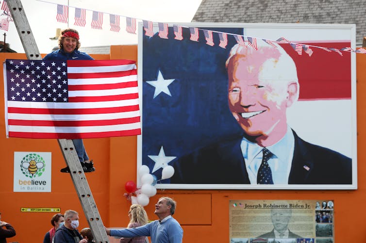 A man holding a US flag on a ladder next to a mural depicting Joe Biden.