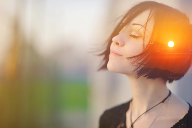 Mujer con el rostro feliz y ojos cerrados y una puesta de sol reflejada en el lateral de su cabeza.