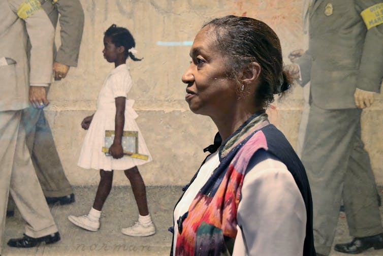 An older woman stands in front of a painting of a young Black girl walking to school