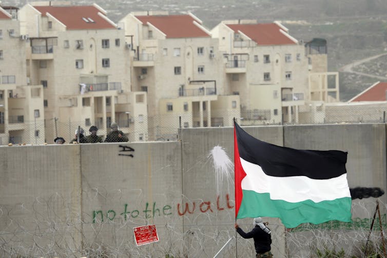 A man waves a Palestinian flag in front of a a wall with graffiti that reads: no to the wall. Israeli soldiers can be seen behind the wall.