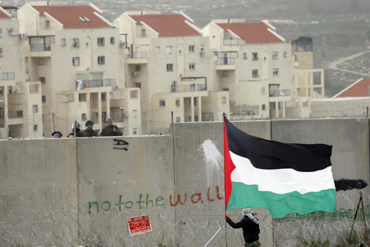 A man waves a Palestinian flag in front of a a wall with graffiti that reads: no to the wall. Israeli soldiers can be seen behind the wall.
