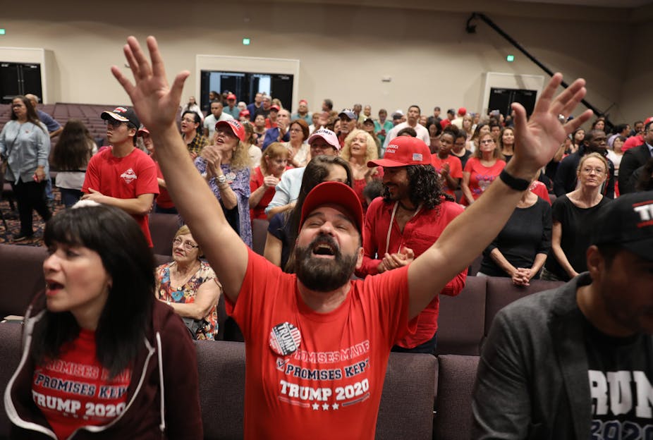 A man wearing a red Trump shirt prays with his hands in the air