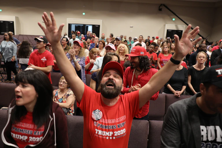 A man wearing a red Trump shirt prays with his hands in the air