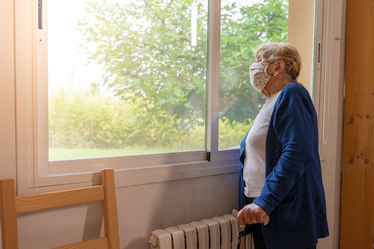 An elderly woman wearing a mask looks out the window.