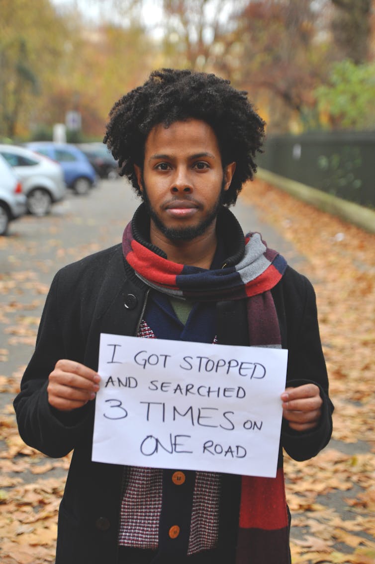 A man holds a sign saying 'I got stopped and searched 3 times on one road'.