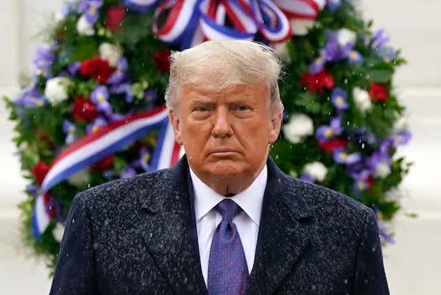 Standing in front of a wreath, Trump has a glum expression on his face as rain falls on him 