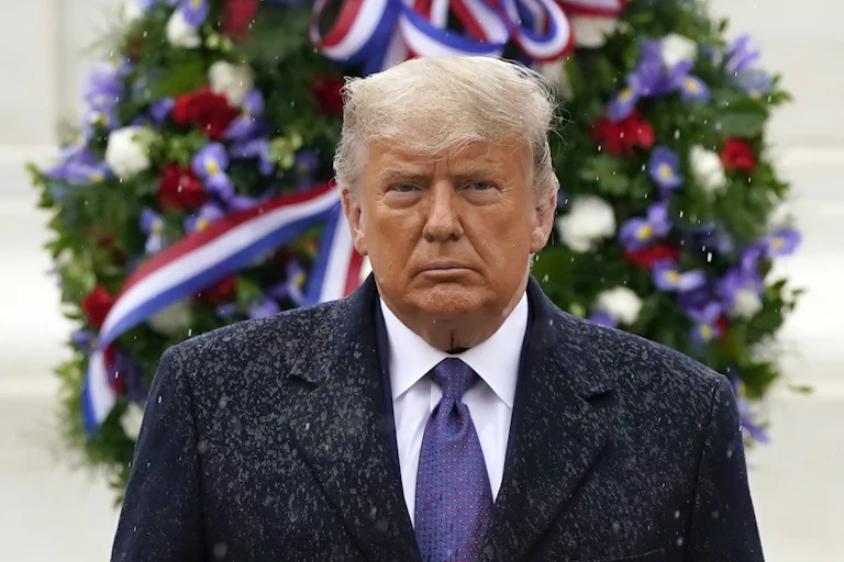 Standing in front of a wreath, Trump has a glum expression on his face as rain falls on him