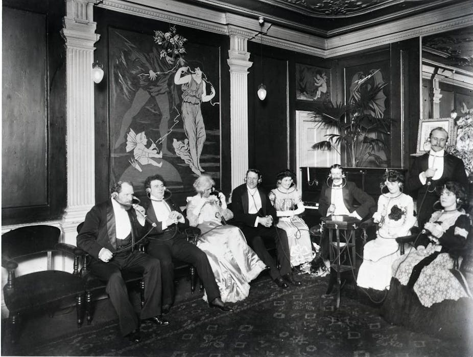 A group of well-dressed men and women from the period listening to infotainment through an Electrophone.