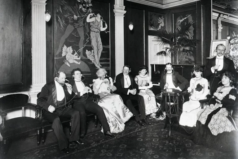 A group of well-dressed men and women from the period listening to infotainment through an Electrophone.
