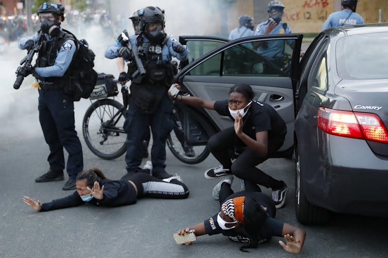 Three women in masks, looking terrified, are on the ground as police carrying assault weapons surround them.
