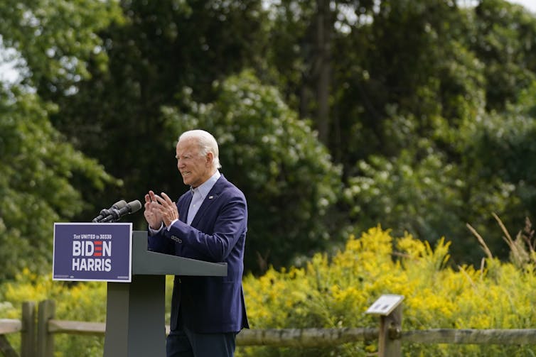 Joe Biden speaking at a lectern outside in front of a stand of trees.